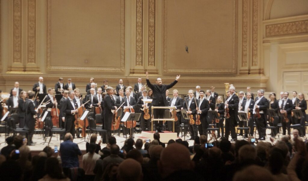 Andris Nelsons with arms raised standing onstage with the Vienna Philharmonic at Carnegie Hall